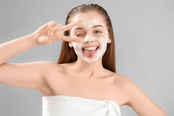 Funny young woman with soap foam on face against grey background