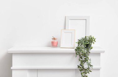 Beyaz duvarın yanındaki şömine rafında boş fotoğraf çerçeveleri, mum ve çiçek var.