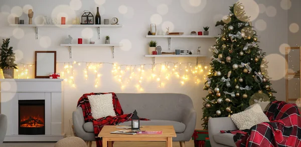 Interior of light living room with glowing garland, Christmas tree, fireplace and sofa