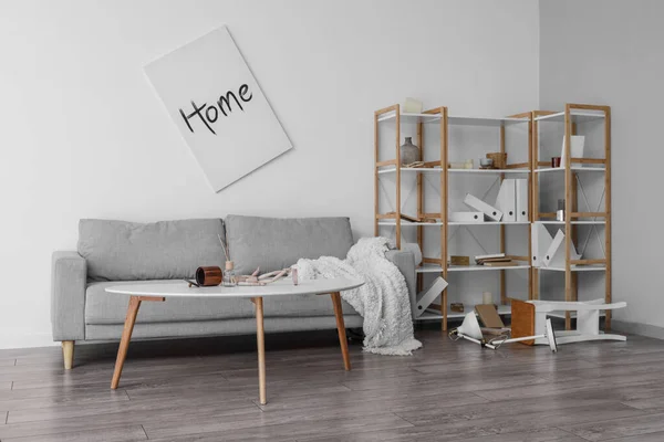 Interior of messy living room with grey sofa, tables and shelving unit