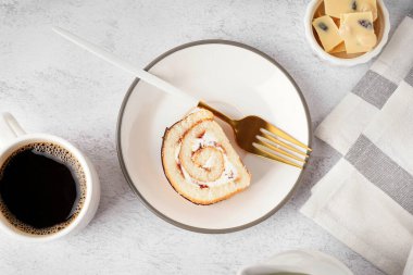 Lezzetli bir parça süngerli kek rulo, çatal, bir fincan kahve ve beyaz masada çikolata.