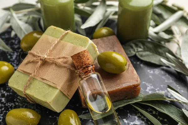 Board with soap bars, bottle of oil, green olives and branches on white background, closeup