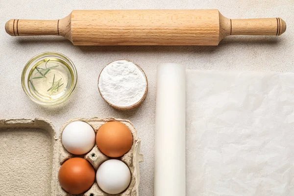 Baking paper with rolling pin, oil, flour and eggs on light background