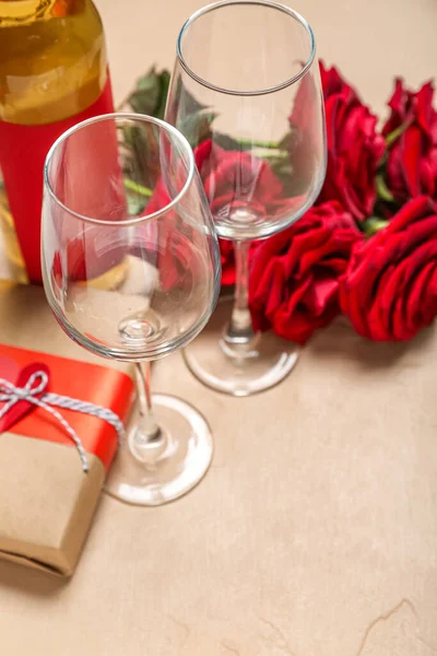 Glasses with gift and roses on beige background, closeup. Valentine\'s Day celebration