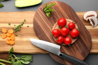 Ahşap kesim tahtası, bıçak ve gri arka planda taze sebzeler.