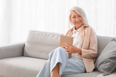 Kitapla evde gri koltukta oturan olgun bir kadın.