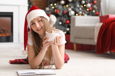 Noel arifesinde evde elinde bir fincan kakao ve defterle pijamalı genç bir kadın.