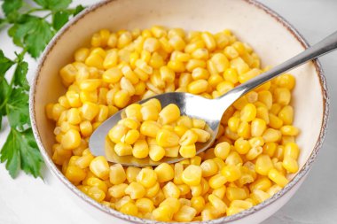 Konserve mısır, kaşık ve maydanozlu kase beyaz masada.