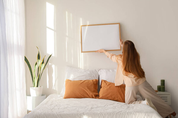 Молодая женщина висит пустой кадр на светлой стене в спальне, вид сзади