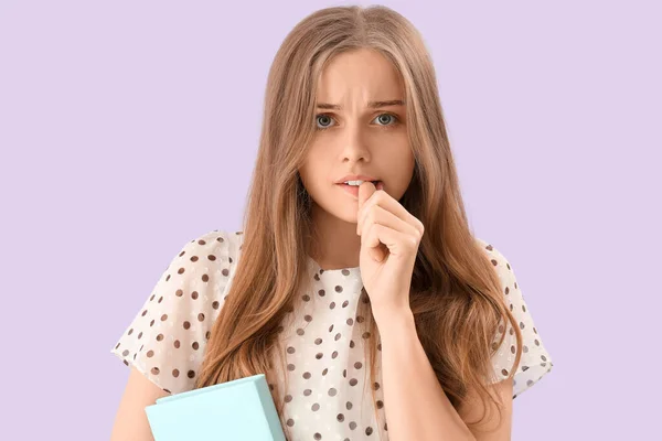 Young woman with book biting nails on lilac background, closeup