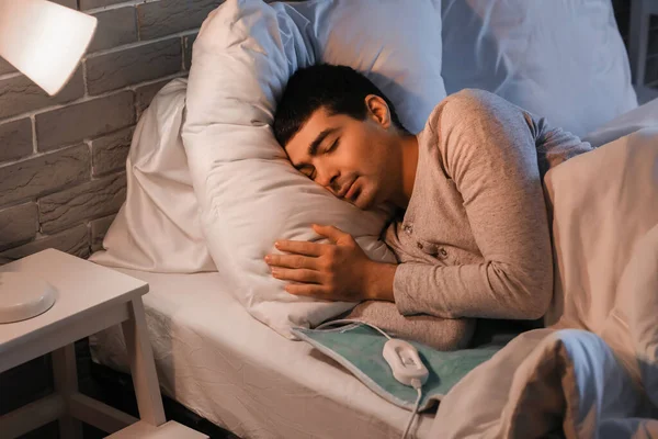 Young man sleeping on electric heating pad in bedroom at night