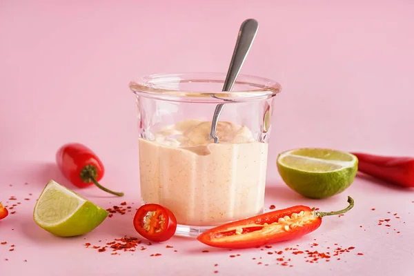 Glass of tasty chipotle sauce, jalapeno peppers and lime on pink background