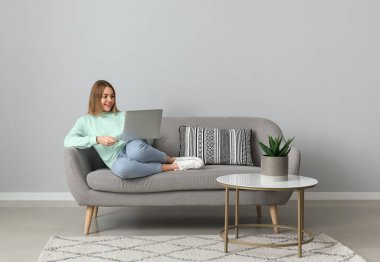 Gri duvarın yanındaki yumuşak koltukta dizüstü bilgisayar kullanan genç bir kadın.