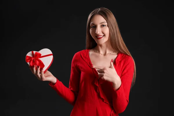 Young woman pointing at gift on black background. Valentine\'s Day celebration