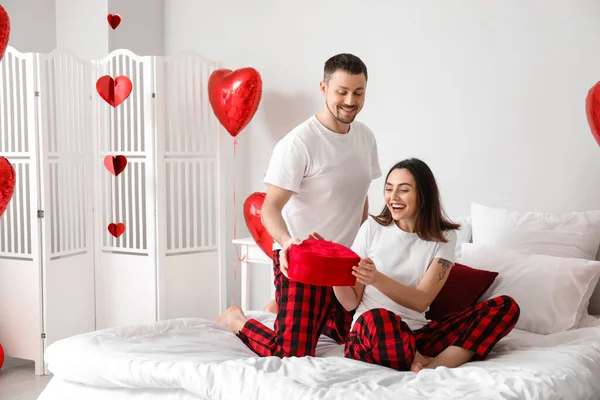 Young man greeting his girlfriend with gift in bedroom on Valentine\'s Day