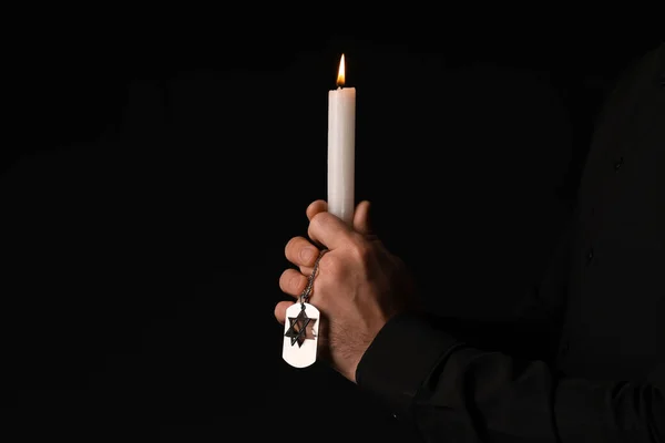 Hands of Jewish man with candle and dog tag honoring victims of Holocaust on dark background