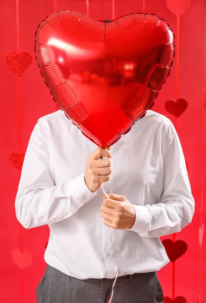 Man hiding face behind heart-shaped balloon on red background. Valentine\'s Day celebration