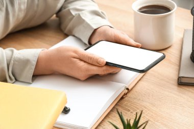 Woman with mobile phone, notebook and cup of coffee at wooden table, closeup
