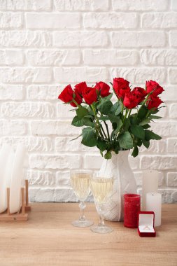 Engagement ring, vase with roses, candles and glasses of champagne on counter near white brick wall
