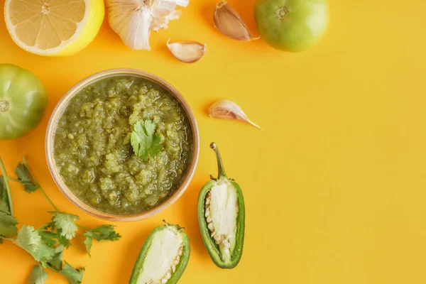 Composition with bowl of tasty green salsa sauce and ingredients on yellow background