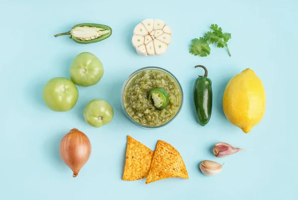 Composition with bowl of tasty green salsa sauce, ingredients and nachos on color background