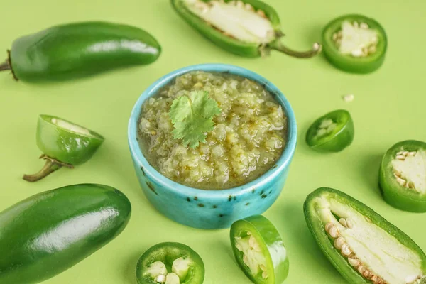 Bowl of tasty green salsa sauce and jalapeno peppers on green background