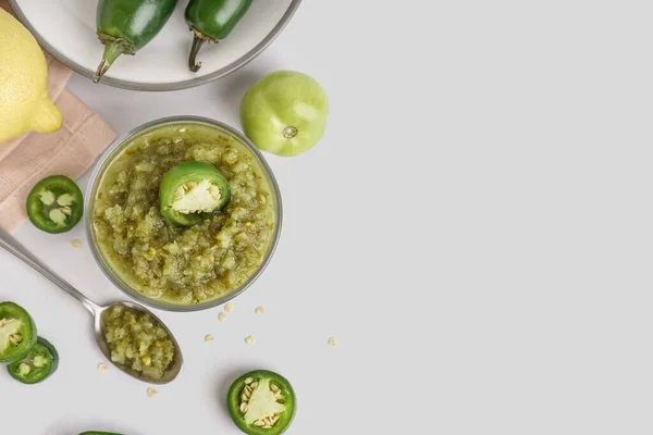 Bowl of tasty green salsa sauce and jalapeno peppers on light background