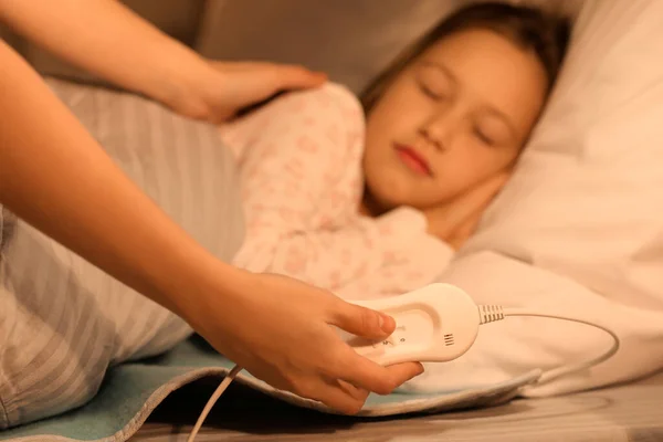 Woman turning on electric heating pad for her little daughter in bedroom at night, closeup