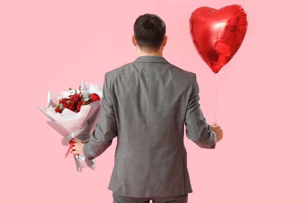 Handsome man with heart-shaped balloon and flowers on pink background, back view. Valentine\'s Day celebration