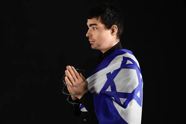 Praying Jewish man with flag of Israel and barbed wire honoring victims of Holocaust on dark background