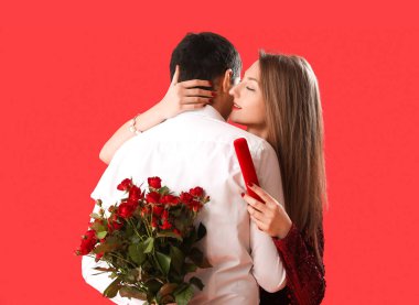 Young woman with gift hugging her boyfriend on red background. Valentine's Day celebration