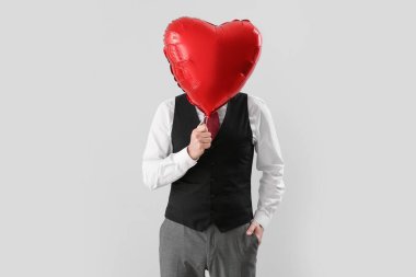 Handsome man hiding face behind heart-shaped balloon on light background. Valentine's Day celebration