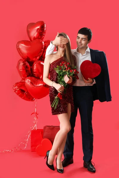 Young couple in love with roses and gift on red background. Valentine\'s Day celebration