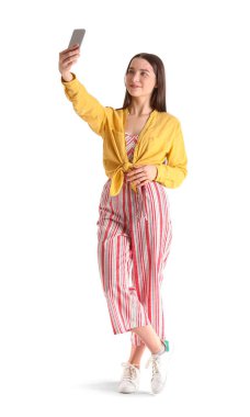 Pretty young woman with smartphone taking selfie against white background