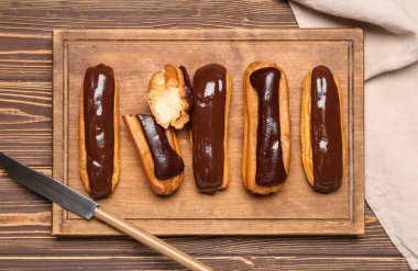Cutting board with tasty chocolate eclairs on wooden background