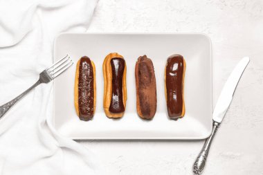 Plate with tasty chocolate eclairs and cutlery on light background