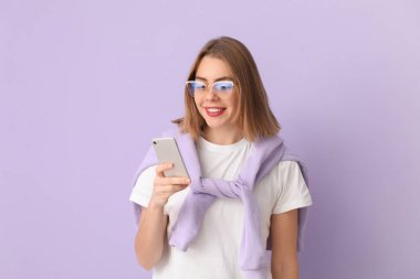Young woman in eyeglasses using mobile phone on lilac background
