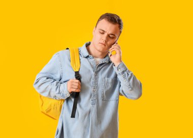 Young man with backpack talking by mobile phone on yellow background