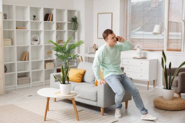 Young man talking by mobile phone near grey couch in living room