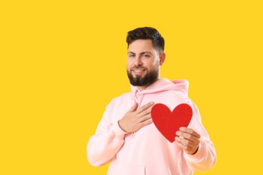 Young bearded man with paper heart for Valentine's Day on yellow background