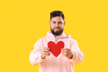 Young bearded man with paper heart for Valentine's Day on yellow background