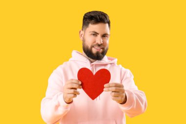 Young bearded man with paper heart for Valentine's Day on yellow background
