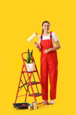Young woman with roller, ladder, houseplant and paint cans on yellow background