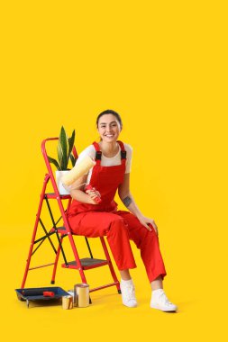 Young woman with ladder, houseplant and paint cans on yellow background
