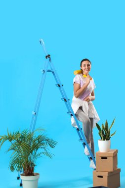 Young woman with paint roller, ladder and moving boxes on blue background