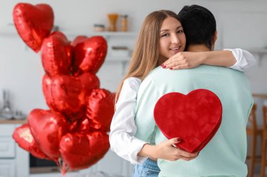 Young woman with gift hugging her boyfriend in kitchen on Valentine's Day