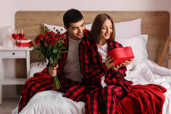 Young couple with roses and gift in bedroom on Valentine\'s Day