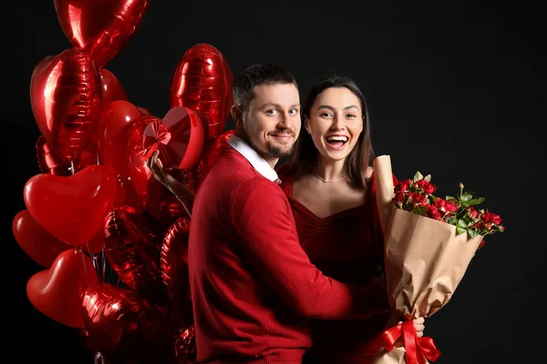 Happy couple in love with roses, gift and balloons on black background. Valentine\'s Day celebration
