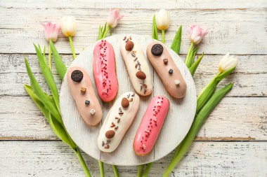 Board with various glazed eclairs and tulips on white wooden background