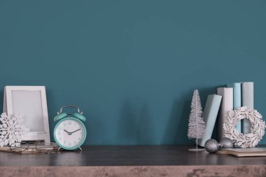 Frame with alarm clock, books and Christmas decor on table near green wall